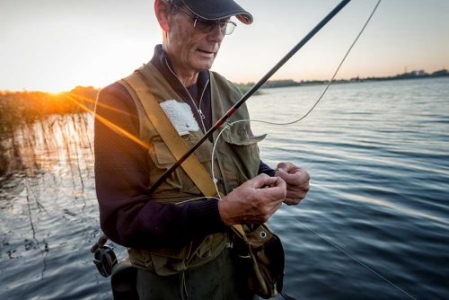 Portrait of a fly fisherman tying a fly on to his line as the sunsets behind him