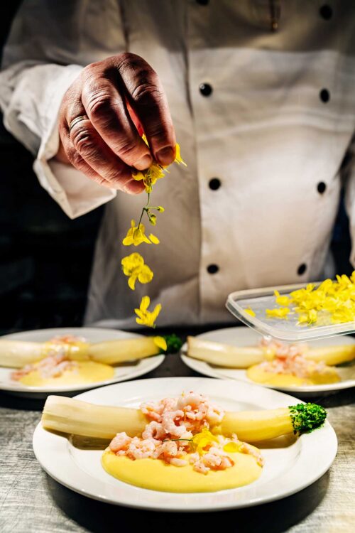 Kokken står i et lyst og veludstyret køkken, hvor han forbereder en lækker ret med hvide asparges, hollandaise sauce og rejer, pyntet med rapsblomster