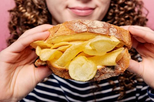 Curly haired girl holding a sandwich filled with potato crisps towards the camera.