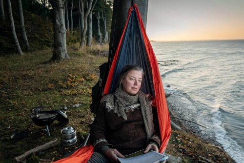 Solens sidste stråler kaster et blødt, gyldent lys over hende og omgivelsen, mens bølgerne blidt ruller ind mod stranden. Hængekøjen svinger let i den milde brise, og kvinden har lukkede øjne med et fredfyldt smil, der udstråler afslapning og lykke. I baggrunden ses silhuetter af palmetræer og en farverig himmel, der skifter fra blå til nuancer af orange og lyserød.