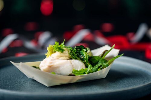 Individual serving of some party food consisting of a small piece of cod topped with marinated seaweed, served with a mustard dressing, wasabi caviar and wood sorrel.