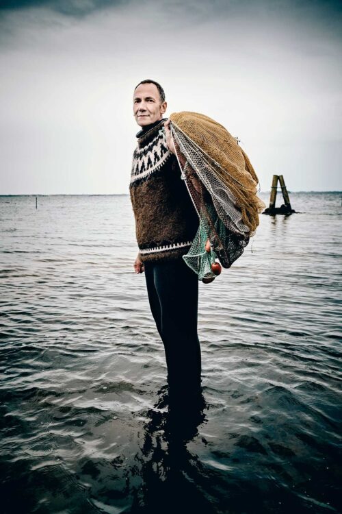 Portrait of a shrimp fisher standing in the water, carrying his nets on his back, looking to camera.