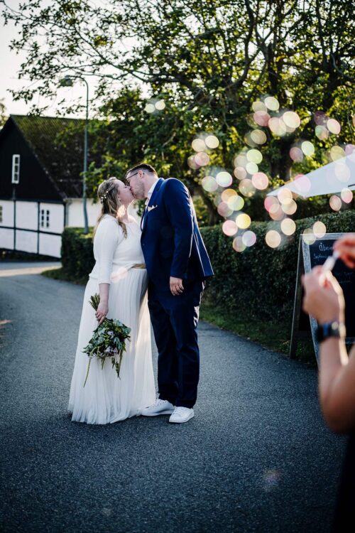 Et nygift par kysser romantisk hinanden, omgivet af blomsterdekorationer og festlig atmosfære. Parret bærer traditionelle bryllupsoutfits, og deres ansigtstræk udstråler lykke og kærlighed.